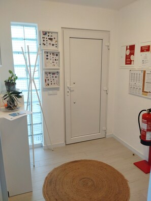 A room with a white door, a window with blinds, a wall-mounted shelf with various items, a red fire extinguisher, and a brown rug on the floor. - Eira da Mila - 2-bedroom house with outdoor area (Coimbra)