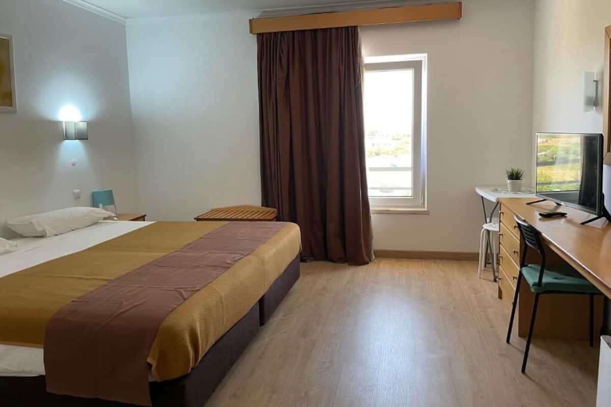 Una habitación de hotel con cama, escritorio, silla, ventana con cortinas y una lámpara en la pared.
