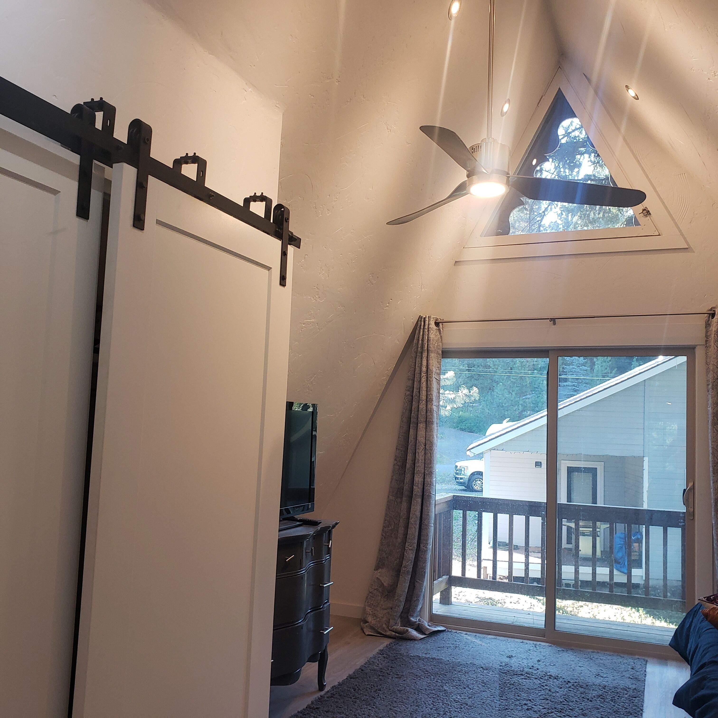 Una habitación con puerta corredera, ventilador de techo, televisor sobre un soporte y vista a una casa desde una ventana.