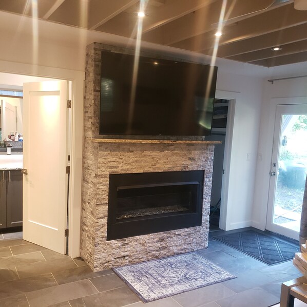 A modern living room with a stone fireplace, a flat-screen TV mounted above it, and a door leading to another room. - Charming 2-bedroom A Frame modern cabin in McCall perfect for a cozy getaway (McCall)