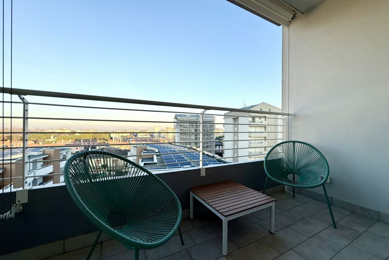 Un balcón con dos sillas verdes, una mesa de madera y vistas a edificios y paneles solares.