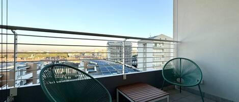 Un balcón con dos sillas verdes, una mesa de madera y vistas a edificios y paneles solares.