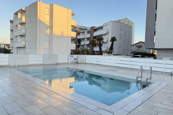 A swimming pool with a tiled deck, surrounded by modern buildings with balconies and a clear sky. - Meit 5b - Carraro Immobiliare Jesolo (Lido di Jesolo)