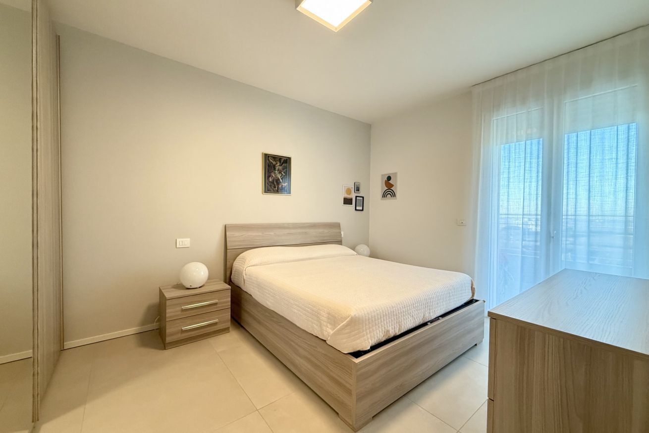 Een moderne slaapkamer met een houten bed, nachtkastjes en een groot raam met dunne gordijnen.