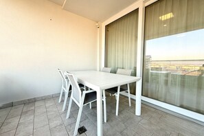 A balcony with a white table and chairs, a large window with curtains, and a view of a cityscape. - Meit 5b - Carraro Immobiliare Jesolo (Lido di Jesolo)