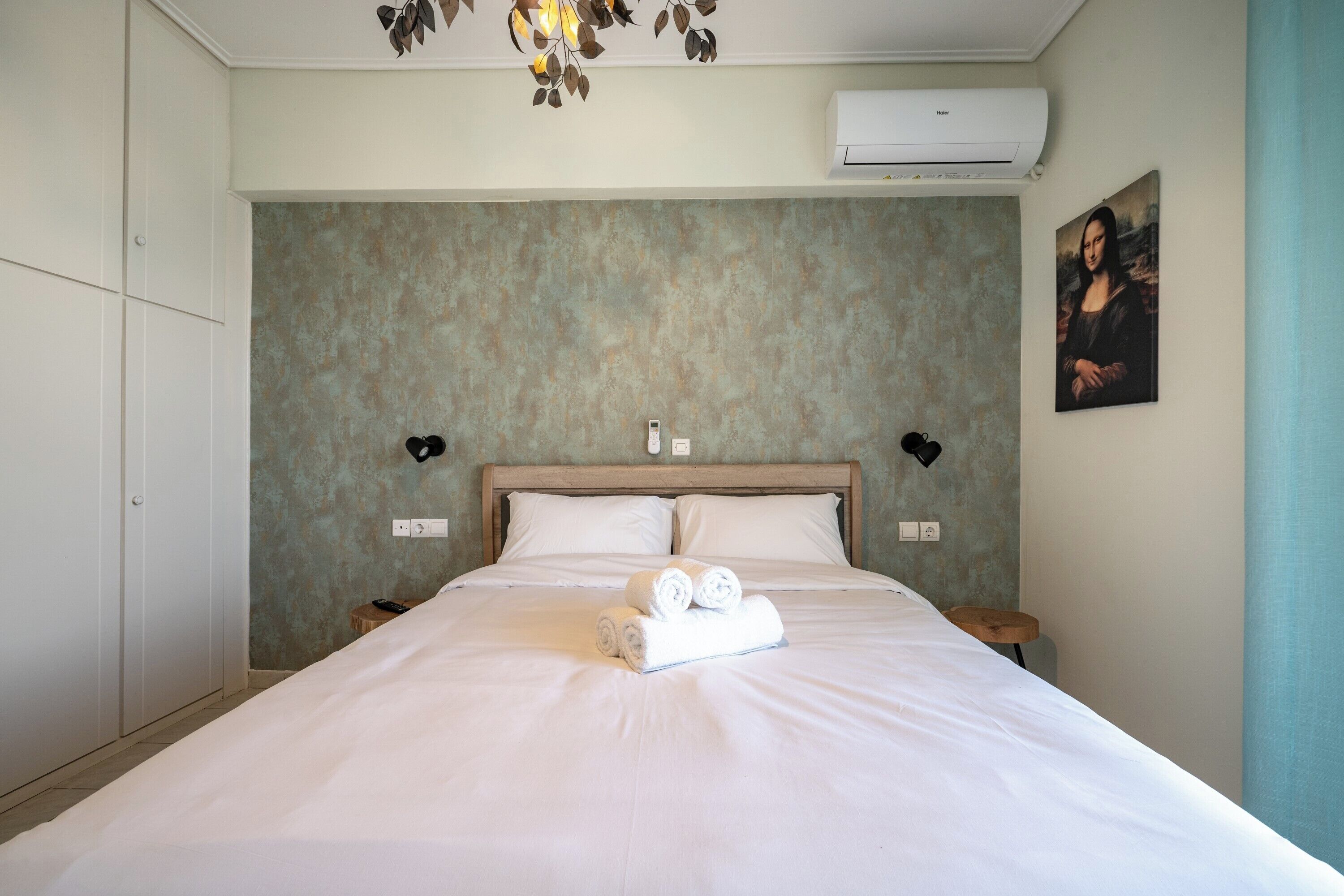 A neatly made bed with rolled towels, a wooden headboard, bedside lamps, and a wall-mounted air conditioner.