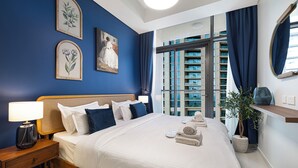 Camera d'albergo con un letto grande, un comodino, una lampada, opere d'arte incorniciate, una pianta in vaso e vista su un edificio dalla finestra.