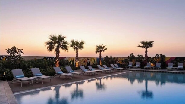 Area piscina con poltronne prendisole e palme sullo sfondo di un cielo al tramonto.