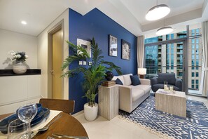 A modern living room with a blue accent wall, a sofa, a coffee table, and a dining area with a table set for two. - Luxury 1BR 5 Min to Burj Khalifa Prime Stay (dubai)