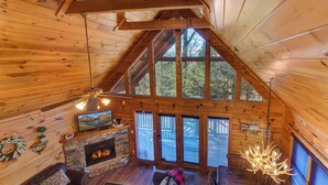 A log cabin interior with a stone fireplace, wooden ceiling beams, a large window, and a chandelier. - Cozy 3-bedroom haven for up to 10 guests. (Gatlinburg)