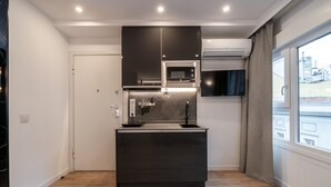 Cozinha compacta com micro-ondas, fogão e pia, uma pequena janela com cortinas e vista para prédios.