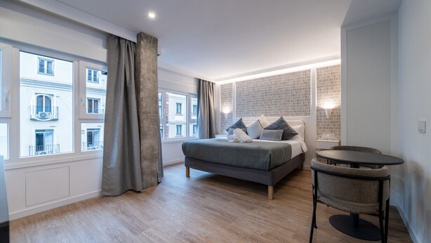 Quarto moderno com cama, mesa redonda e vista para edifícios através de grandes janelas.