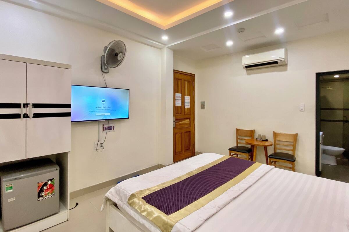 A hotel room with a bed, a TV mounted on the wall, a fan, a small table with chairs, and an air conditioning unit.