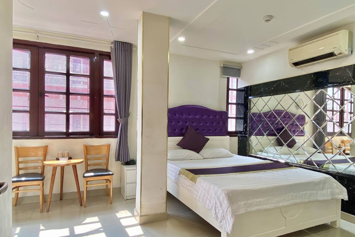 A hotel room with a bed, a small table with chairs, a window with curtains, and an air conditioning unit.