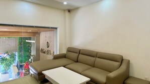 Un soggiorno moderno con un divano modulare color beige, un tavolino da caffè bianco e una porta vetrata che dà su un balcone con piante e bandiere rosse.