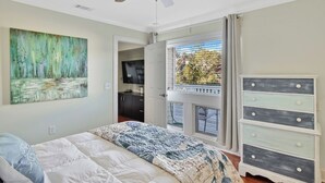 A bedroom with a bed, a ceiling fan, a painting, a dresser, and a window with a view of trees. - 2 bedroom 2 bath condo with deck overlooking lagoon. (Saint Simons Island)