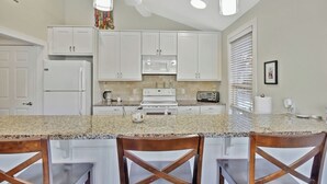 A kitchen with white cabinets, a granite countertop, a stove, a microwave, and a toaster. There are three wooden chairs at the counter. - 2 bedroom 2 bath condo with deck overlooking lagoon. (Saint Simons Island)