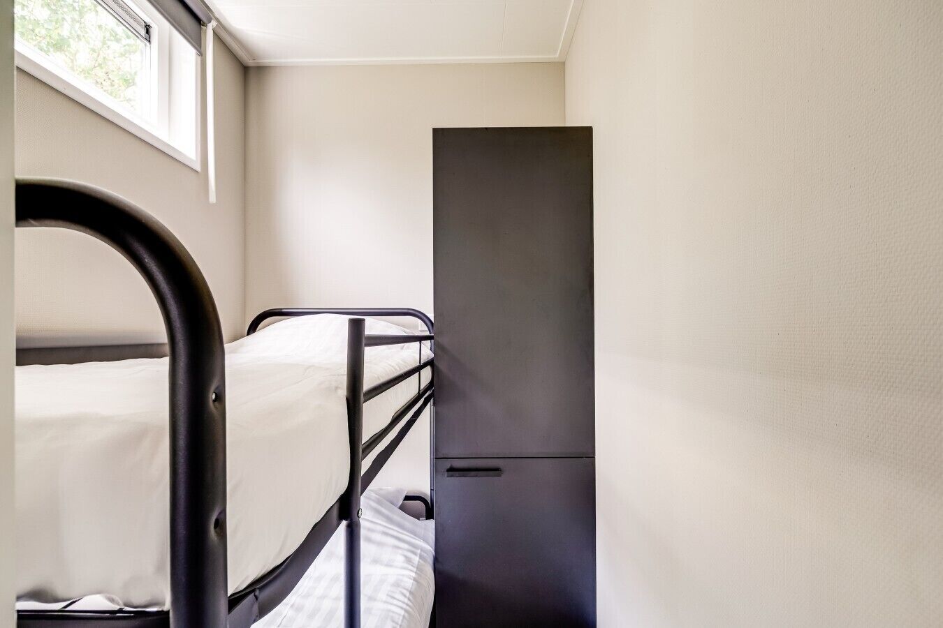 Un lit simple avec un cadre en métal noir, une porte et une fenêtre donnant sur un paysage verdoyant.