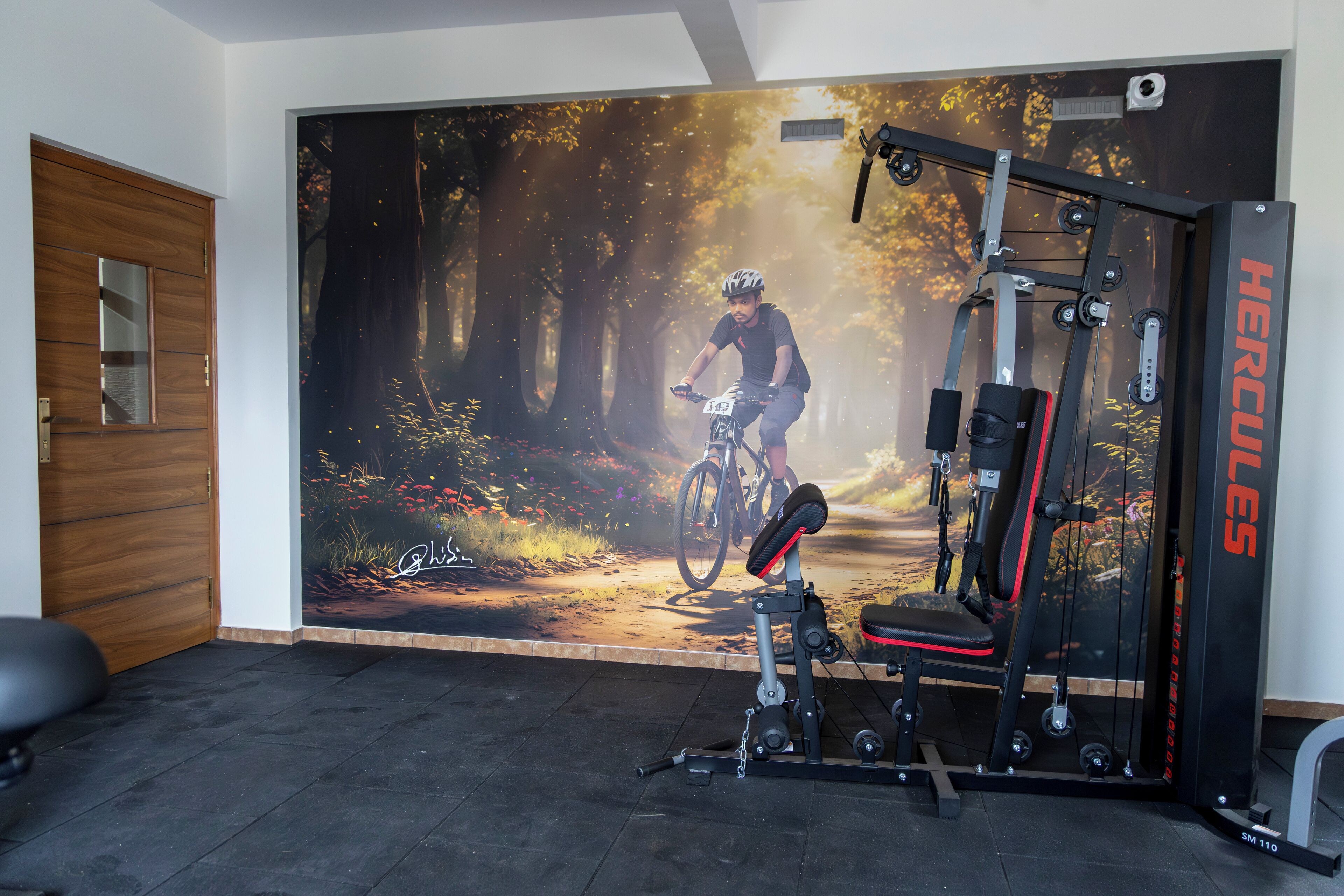 Un gimnasio con un gran mural de un ciclista en un sendero de montaña, una máquina de pesas Hercules y una puerta de madera.