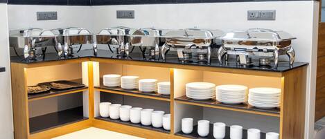 A buffet setup with multiple serving stations, including covered chafing dishes and neatly stacked plates and bowls.