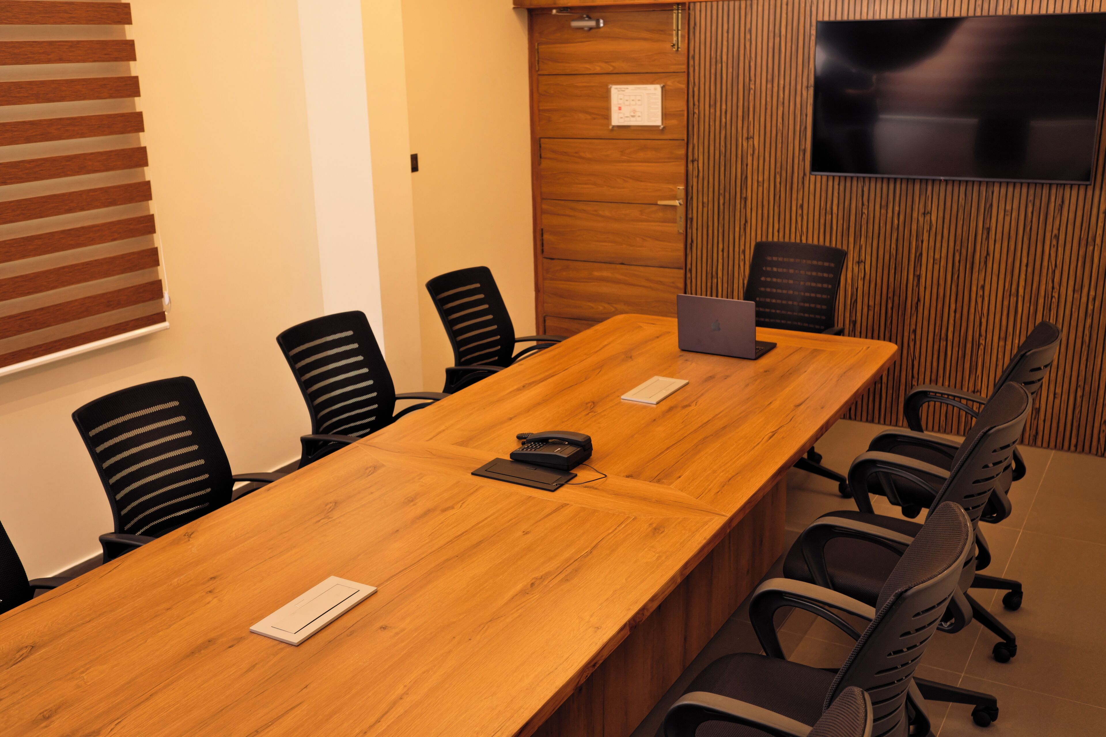 A conference room with a long wooden table, black office chairs, a laptop, a telephone, and a TV mounted on the wall.