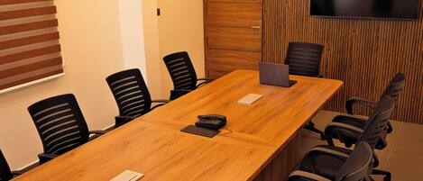 A conference room with a long wooden table, black office chairs, a laptop, a telephone, and a TV mounted on the wall.