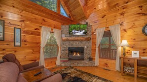 Un interior de cabaña acogedor con chimenea de piedra, paredes de madera, un televisor montado encima y una zona de estar cómoda con sofá y una mesa de centro.