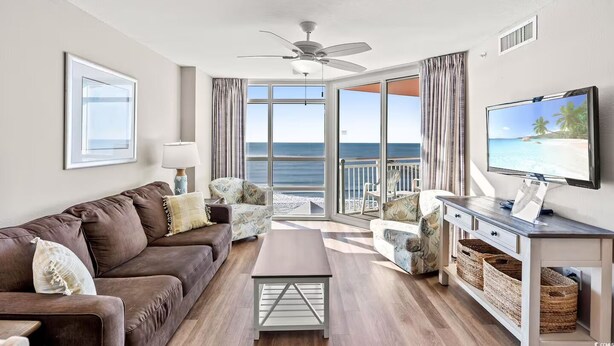 A living room with a brown sofa, a coffee table, and a TV on a stand. There are two chairs and a window with a view of the ocean. - Cozy 2-bedroom condo with AC in peaceful North Myrtle Beach with amazing view (North Myrtle Beach)