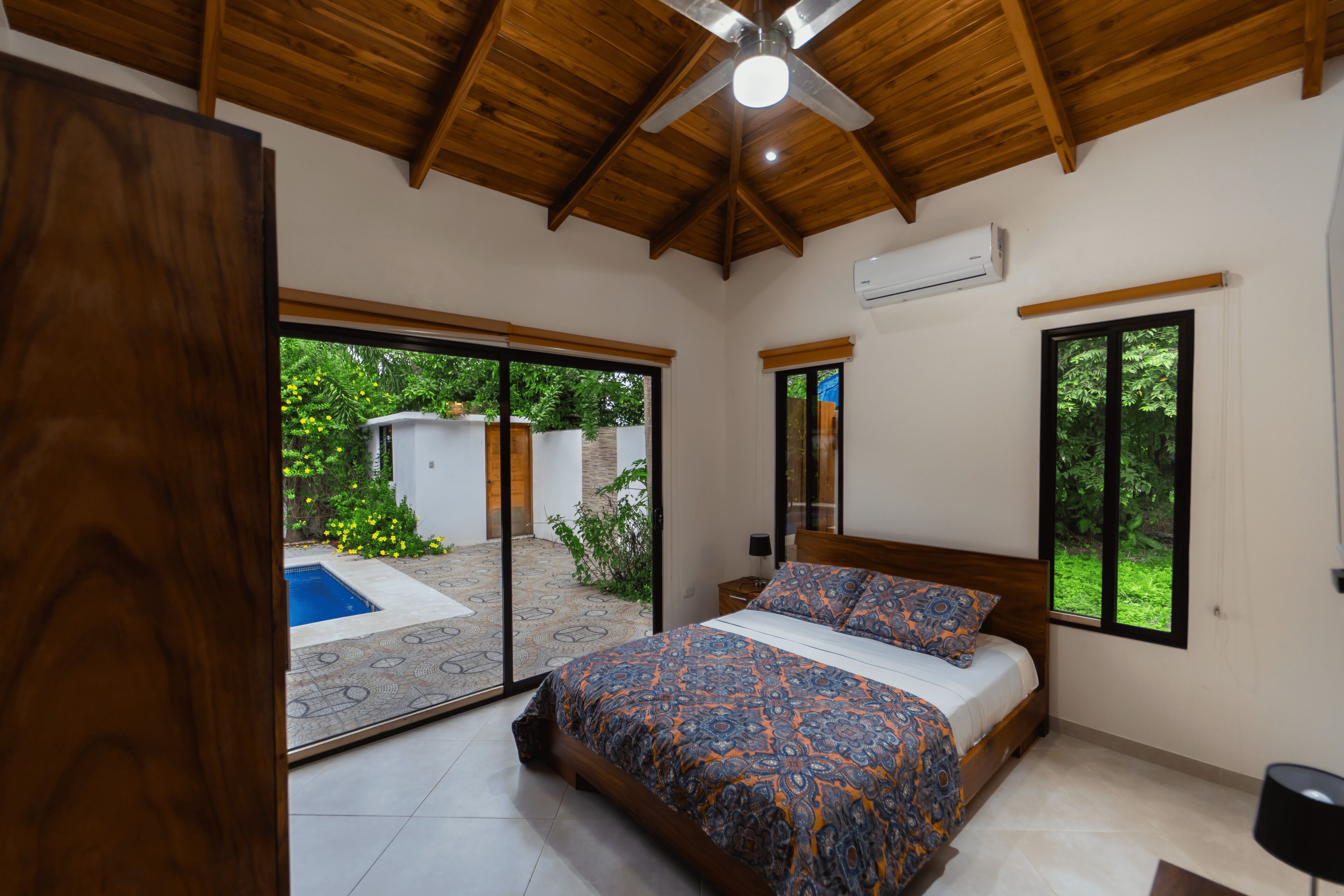 Une chambre dotée d’un grand lit, de poutres apparentes en bois au plafond et offrant une vue sur une piscine et un jardin.