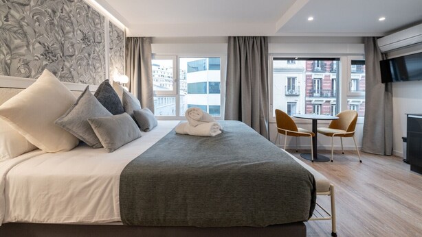 A modern hotel room with a large bed, a small table with two chairs, a flat-screen TV, and a view of the city through large windows. - 302 — SB20 Boutique Studios | 1BR | 1BA | W/K (Madrid)