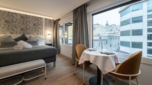 A modern hotel room with a bed, a small table with chairs, and a view of the city through large windows. - 302 — SB20 Boutique Studios | 1BR | 1BA | W/K (Madrid)