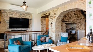 Ein Wohnzimmer mit Steinwänden, einem Flachbildfernseher, blauen Sofas, einem Holztisch und einem Steingewölbe, das in eine Küche führt.