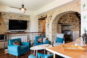 A living room with stone walls, a flat-screen TV, blue sofas, a wooden table, and a stone archway leading to a kitchen. - Beachfront House with Hot Tub, Walk to Everything! (Panormos in Rethymno)