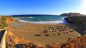 Ein Strand mit Sonnenschirmen und Liegestühlen, felsigen Klippen und blauem Himmel.