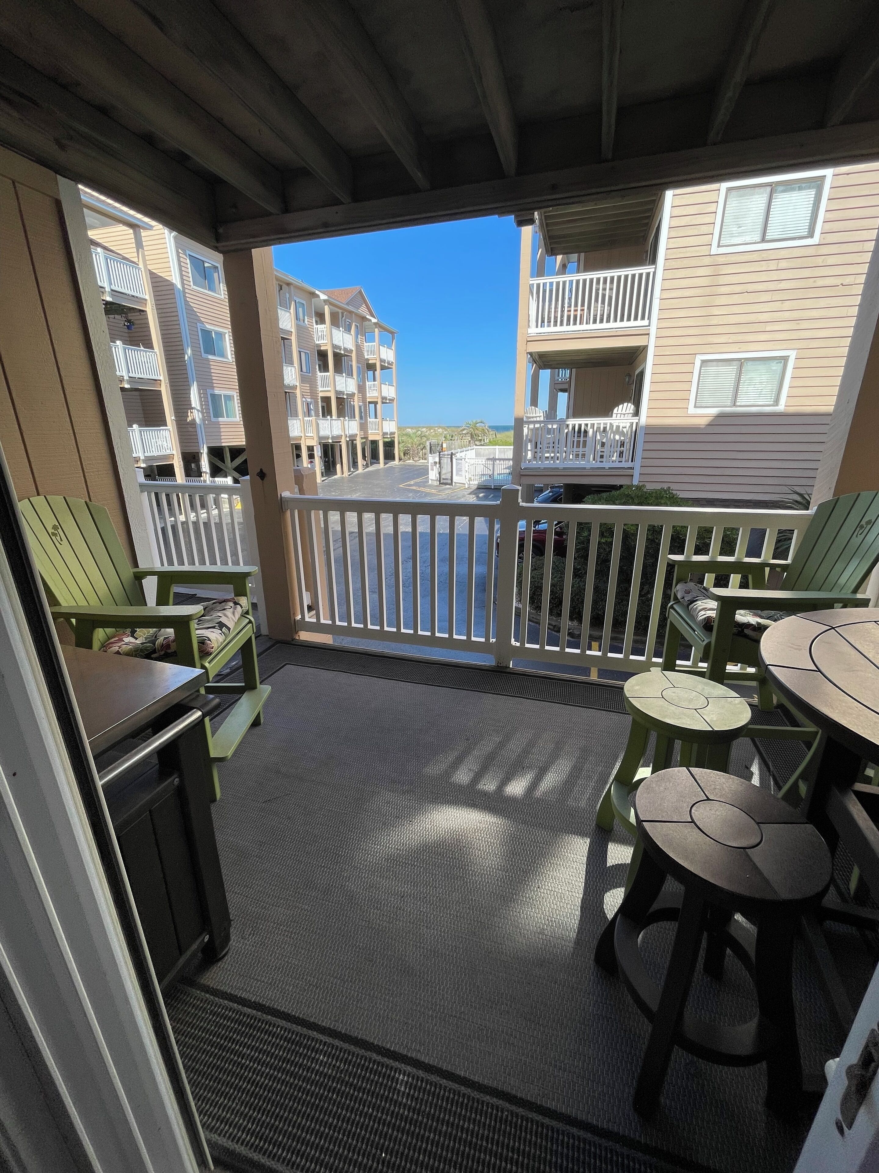 A covered outdoor area with a patio, green chairs, a table, and a view of a building with balconies and a body of water in the background.