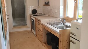 Eine kleine Küche mit Spüle, Herd und Kühlschrank. Ein Fenster sorgt für natürliches Licht und eine Tür führt zu einem Duschbereich.