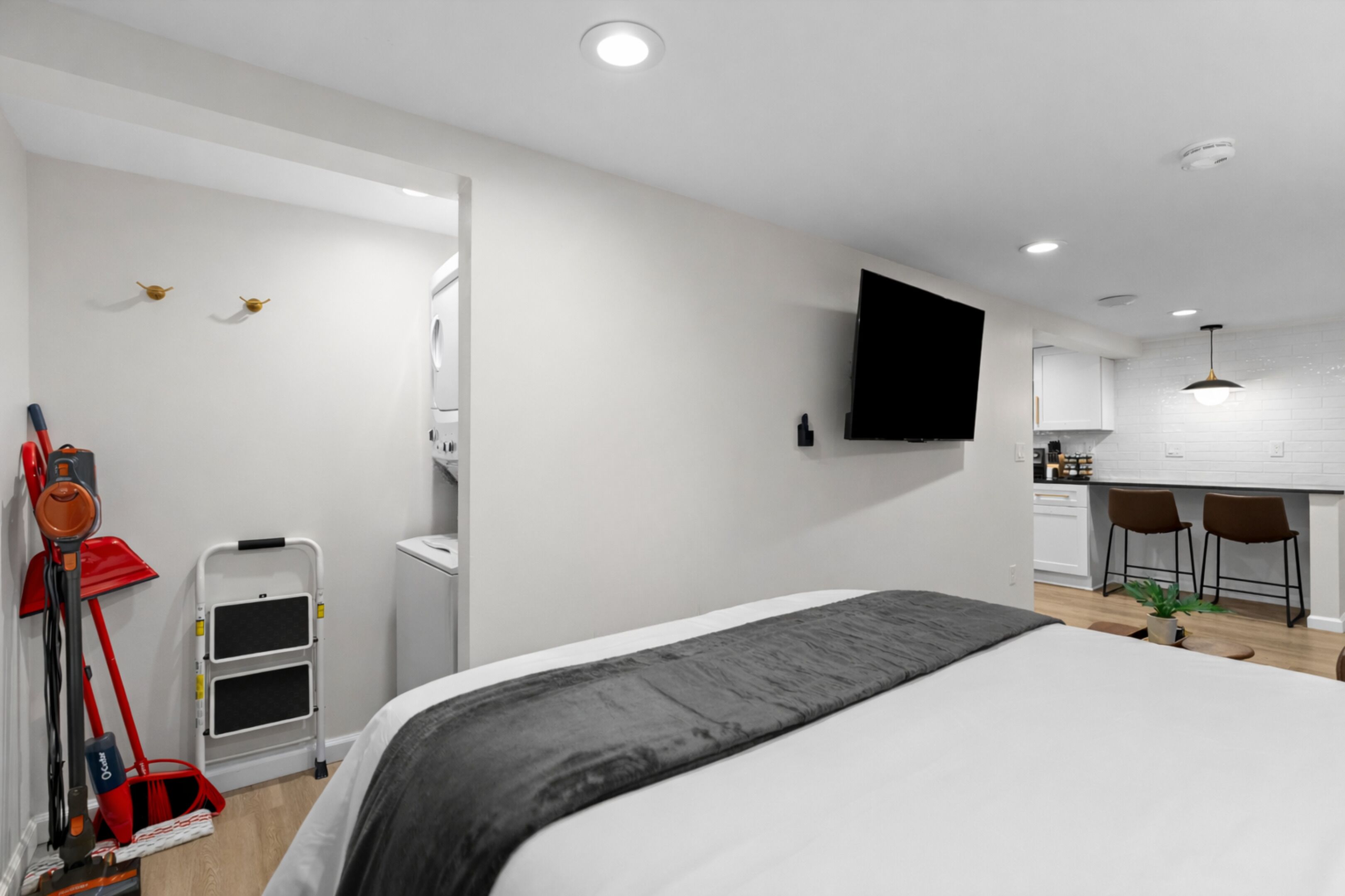Ett modernt hotellrum med en stor säng, en platt-TV som är monterad på väggen, en liten köksdel med barstolar och en städvagn.