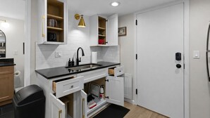Una cocina moderna con armarios blancos, encimera negra y un lavamanos integrado. Hay repisas abiertas con varios objetos y un reloj en la pared.