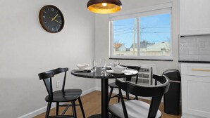 Una cocina moderna con armarios blancos, una mesa redonda de comedor con sillas negras, un reloj de pared y una ventana con vista a casas.