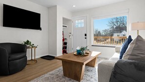 Una sala moderna con una mesa de centro de madera, un sillón de cuero negro, un sofá gris y un ventanal con vistas a una terraza de madera y árboles.