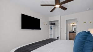 A bedroom with a bed, a ceiling fan, a TV, a wardrobe, and a bathroom visible through an open door. - Charming 1BR Getaway | Eastpointe (Eastpointe)