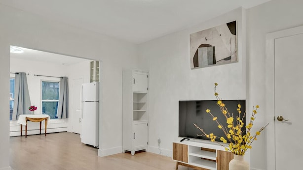 Un soggiorno moderno con una TV a schermo piatto, un mobiletto bianco, un'area pranzo con tavolo e sedie e un'ampia finestra con tende.