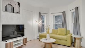 Un soggiorno moderno con televisione a schermo piatto, un divano giallo, un vaso bianco con fiori gialli e un tavolino rotondo.