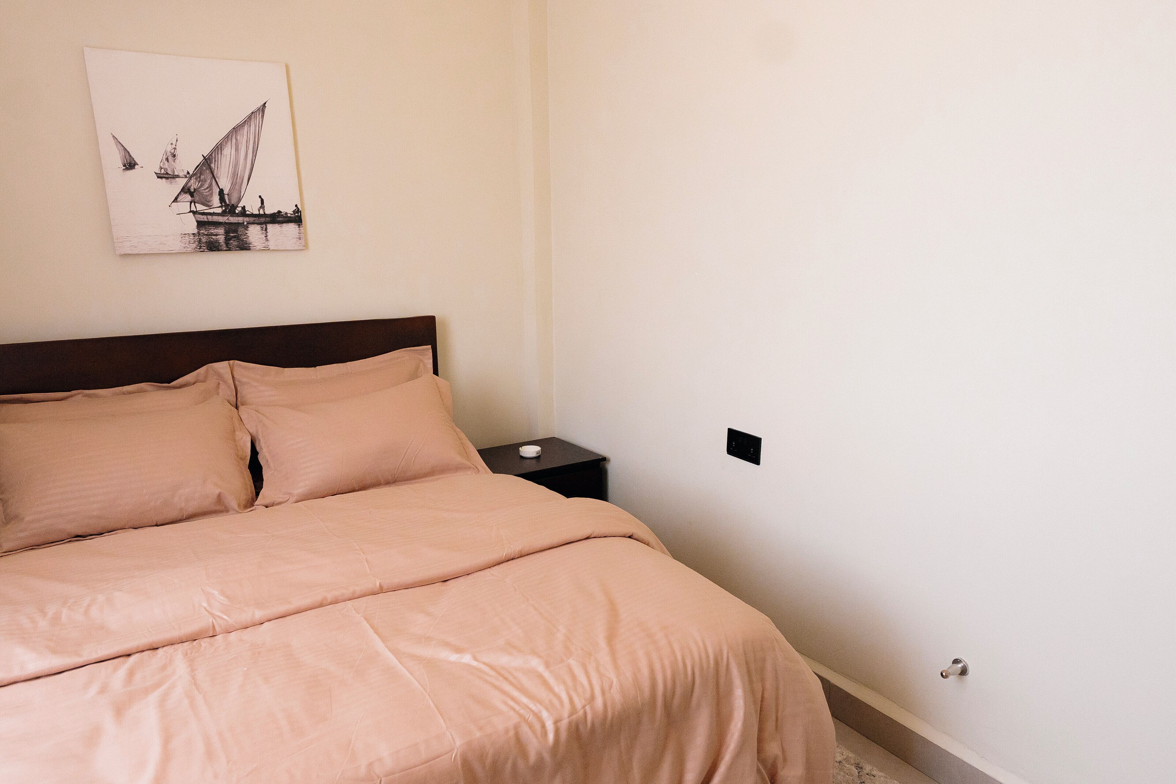A neatly made bed with a headboard, a bedside table with a lamp, and a wall-mounted artwork depicting a sailboat.