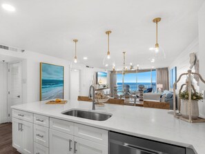 A modern kitchen with a large island, stainless steel sink, and white cabinetry. There is a view of the ocean through large windows. - On the Beach with Balcony, Pool, and Sunsets (Miramar Beach)