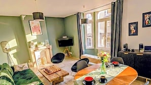 A living room with a green sofa, a wooden coffee table, a dining table with chairs, a TV, and a window with curtains. - L'Écrin d'Opale: Historic Suite 60m² • King Size Bed • Old Town (Boulogne-sur-Mer)