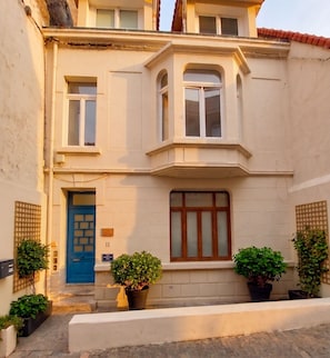 A two-story building with a blue door, a large window with a wooden frame, and potted plants in front.