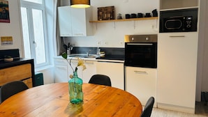 A modern kitchen with white cabinets, a wooden dining table, and a hanging pendant light. - L'Écrin d'Opale: Historic Suite 60m² • King Size Bed • Old Town (Boulogne-sur-Mer)
