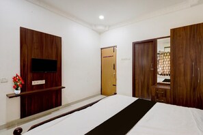 A hotel room with a bed, a TV mounted on a wooden panel, a wooden wardrobe, and a door leading to another room.
