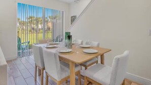 A dining area with a wooden table set for four, white chairs, and a view of the outdoors through large windows. - Cozy villa in Margaritaville Orlando, near Disney, SeaWorld, Universal (Kissimmee)